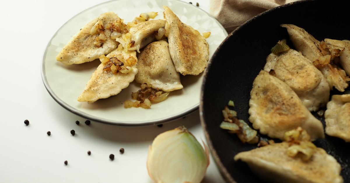 Talerz z pierogami podsmażonymi na złoto z cebulką obok patelni z kolejną partią pierogów oraz cebulą i ziarnami pieprzu na blacie.