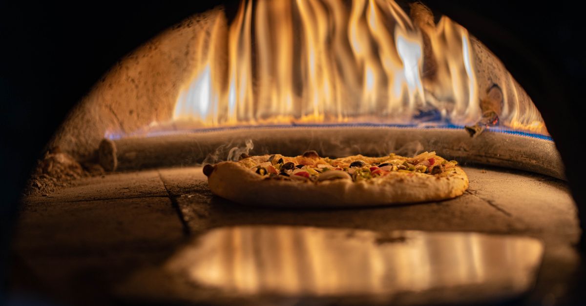 Pizza z serem i warzywami piecze się na kamiennej płycie w rozgrzanym piecu opalanym drewnem, z widocznym płomieniem w tle.