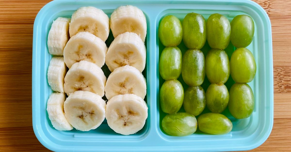 Pokrojone plastry banana i winogrona ułożone w turkusowym pojemniku na drewnianym blacie.