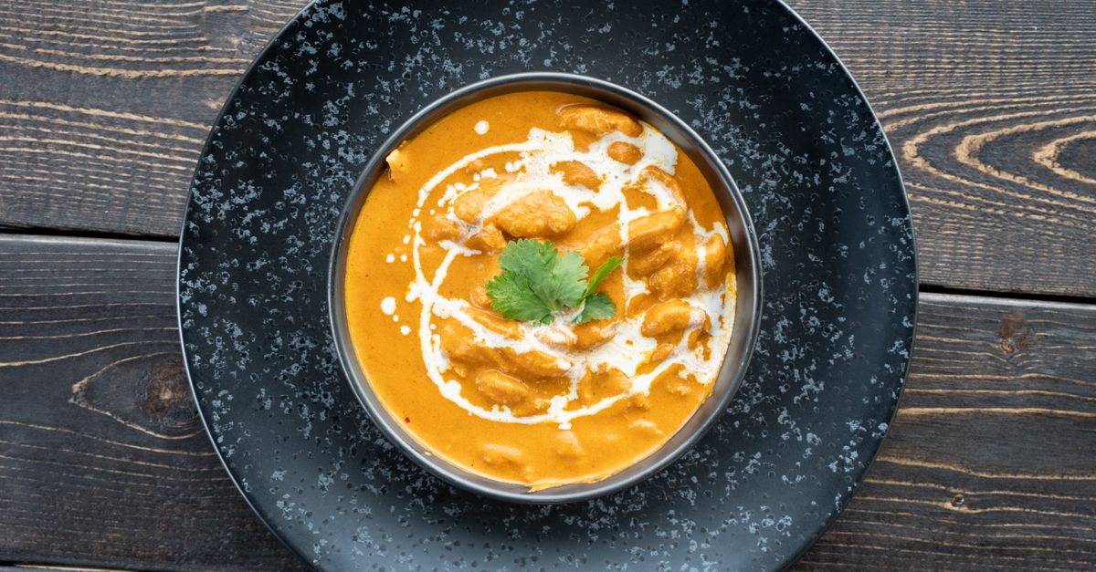 Miska aromatycznego pomarańczowego curry z kawałkami kurczaka, polana śmietaną i udekorowana listkiem kolendry, podana na czarnym talerzu na drewnianym stole.