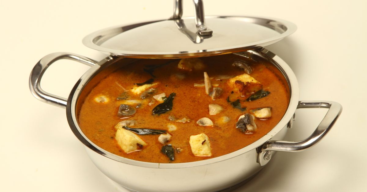 Metalowy garnek z pokrywką wypełniony pikantnym curry z kawałkami sera paneer, grzybami i liśćmi curry w czerwonym sosie.