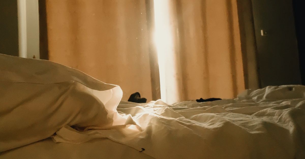 Promienie słonecznego światła przebijające się przez widoczne w powietrzu drobinki kurzu nad łóżkiem z pościelą, na tle zasłoniętego zasłonami okna.